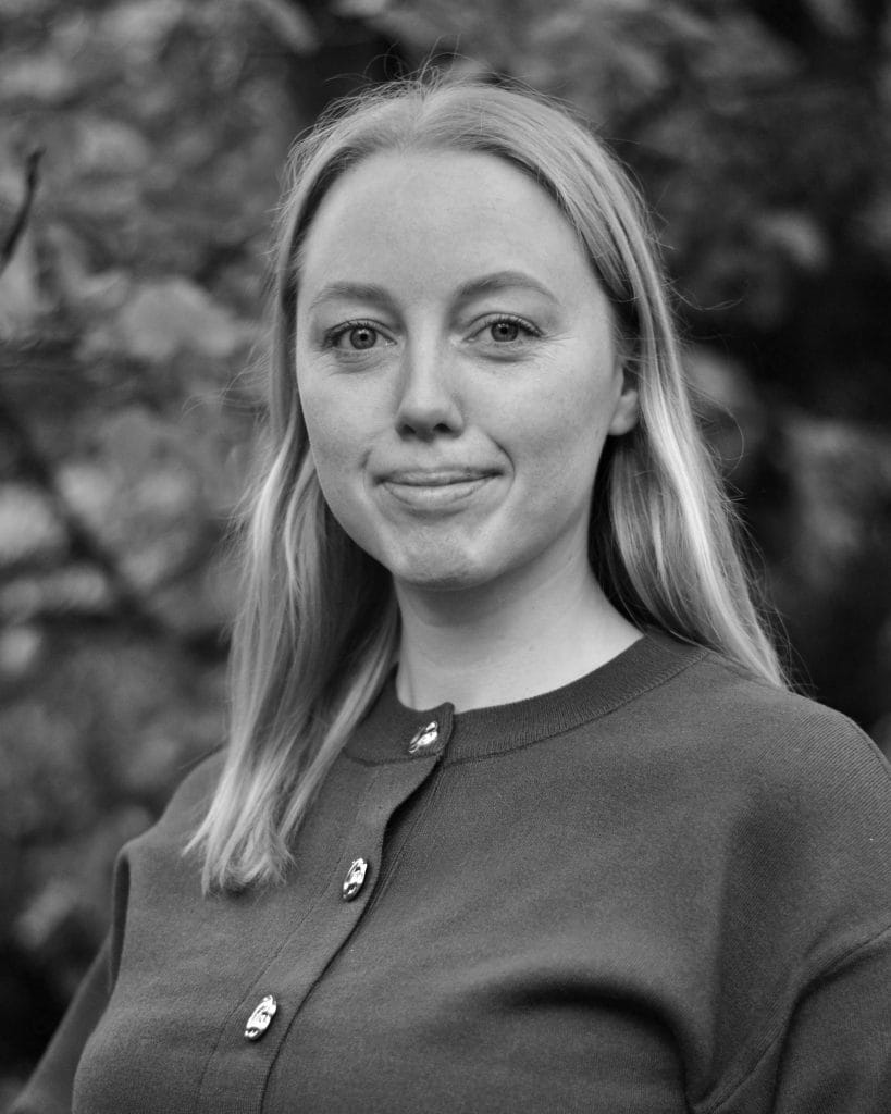 svart-hvitt bilde av kvinne med halvlangt lyst hår, smiler til kamera.